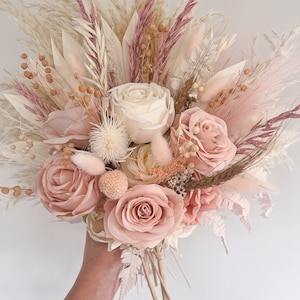 May include: A bridal bouquet featuring a mix of dried and faux flowers in soft pink, cream, and beige tones. The arrangement includes roses, pampas grass, and other textured elements, creating a romantic and natural aesthetic.