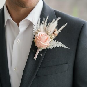 May include: A dark gray suit jacket with a white dress shirt and a pink rose boutonniere. The boutonniere includes a pink rose, white fern, and dried flowers. The jacket has a pocket on the left side.