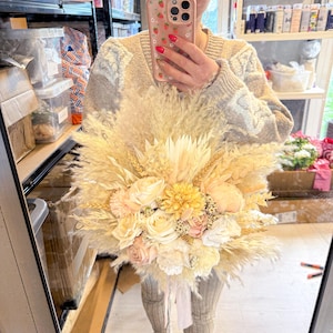 May include: A large, dried floral bouquet featuring pampas grass, wheat, and various flowers in cream, peach, and yellow hues. The bouquet is tied with a white ribbon. The arrangement is held in front of a mirror.