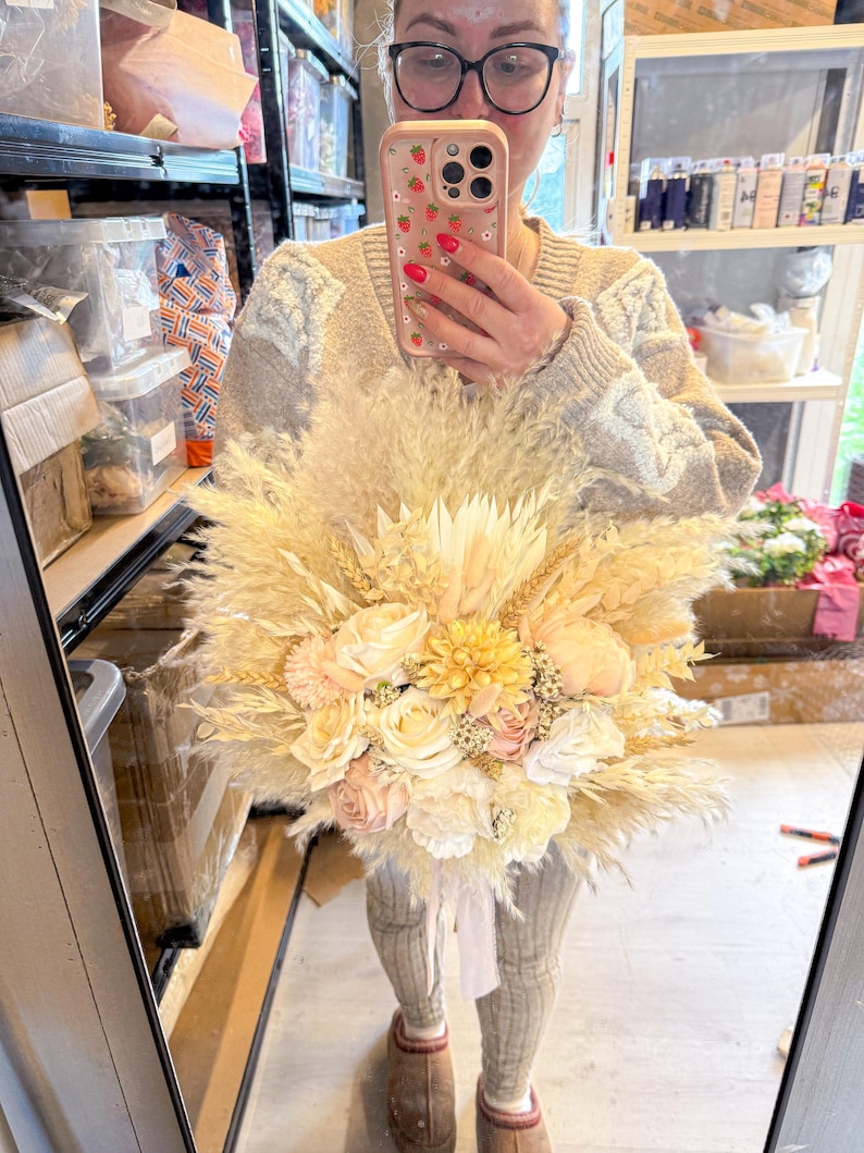May include: A large, dried floral bouquet featuring pampas grass, wheat, and various flowers in cream, peach, and yellow hues. The bouquet is tied with a white ribbon. The arrangement is held in front of a mirror.
