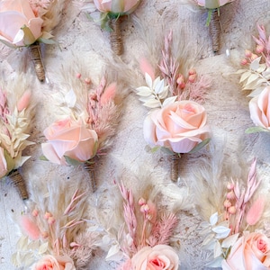 May include: A collection of blush-colored rose boutonnieres with dried pampas grass and other delicate floral accents. The roses have a soft, peachy hue, and the stems are wrapped in twine. These boutonnieres are perfect for weddings or special events.