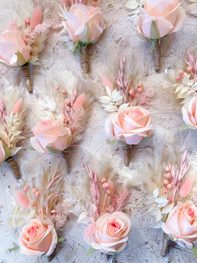 May include: A collection of blush-colored rose boutonnieres with dried pampas grass and other delicate floral accents. The roses have a soft, peachy hue, and the stems are wrapped in twine. These boutonnieres are perfect for weddings or special events.