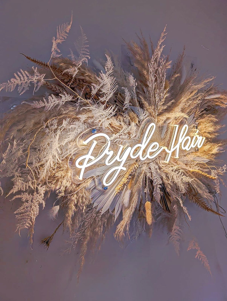 May include: A decorative arrangement of dried grasses and foliage, including pampas grass and ferns, in shades of beige and brown. A neon sign that reads "Dryde Hair" is centered within the arrangement, illuminated in white.