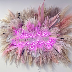 May include: A pink neon sign with the words "You're amazing" surrounded by dried pampas grass in shades of pink, white, and beige.