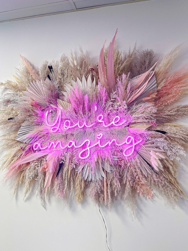May include: A pink neon sign with the words "You're amazing" surrounded by dried pampas grass in shades of pink, white, and beige.
