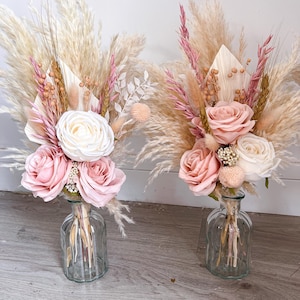 May include: Two floral arrangements in clear glass vases. Each arrangement features a mix of pink roses, white roses, and dried pampas grass. The vases have a textured, ribbed design, and the overall aesthetic is soft and romantic.