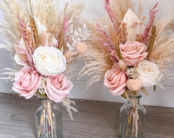Op de afbeelding: Twee bloemstukken in heldere glazen vazen. Elk arrangement bevat roze en witte rozen en gedroogd pampasgras. De vazen hebben een gestructureerd, geribbeld ontwerp, wat zorgt voor een zachte, romantische uitstraling.