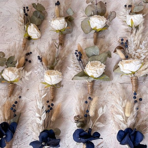 May include: Twelve buttonholes with a rustic, boho theme.  Six feature white roses, dried grasses, and eucalyptus leaves.  Six feature navy blue flowers, dried grasses, and eucalyptus leaves.