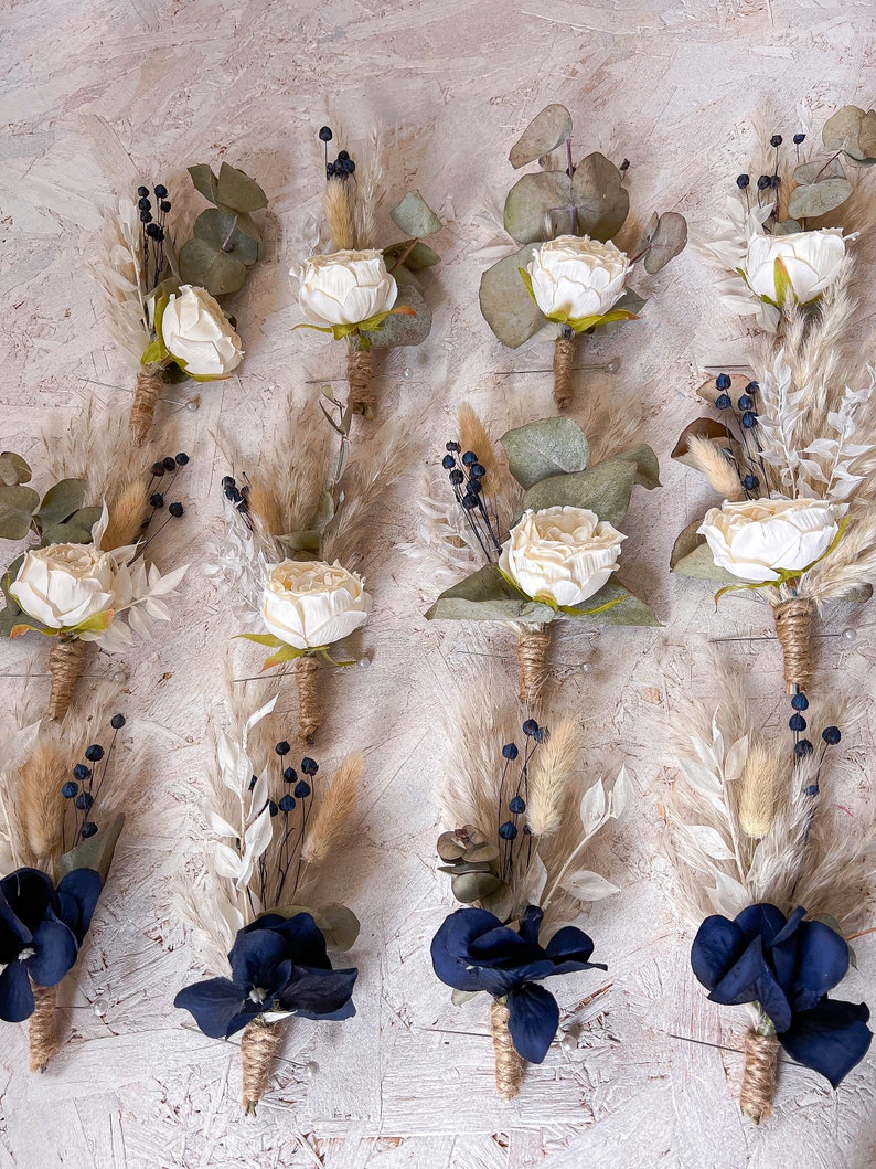 May include: Twelve buttonholes with a rustic, boho theme.  Six feature white roses, dried grasses, and eucalyptus leaves.  Six feature navy blue flowers, dried grasses, and eucalyptus leaves.