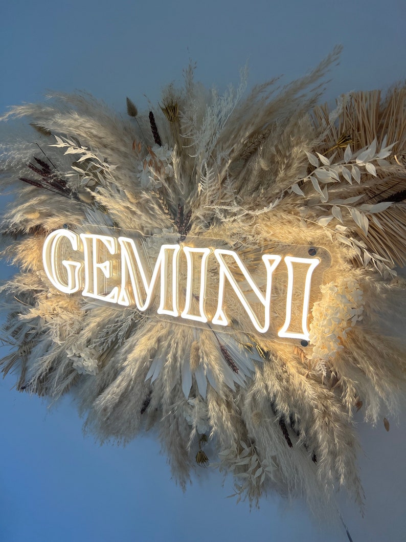 May include: A white neon sign with the word "GEMINI" in a cursive font, surrounded by a wreath of dried pampas grass and other dried flowers.