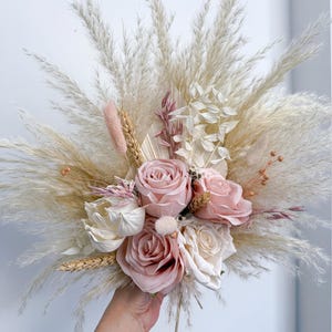 May include: A bridesmaid-sized bouquet featuring a mix of dried pampas grass, wheat, and faux roses in shades of pink and cream. The bouquet includes the text "BRIDESMAID SIZE".