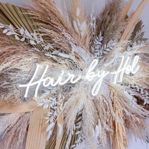 May include: A decorative wall hanging featuring dried pampas grass, palm leaves, and white floral accents. A neon sign that reads "Hair by Hol" is mounted in the center of the arrangement. The color palette includes beige, cream, and white.