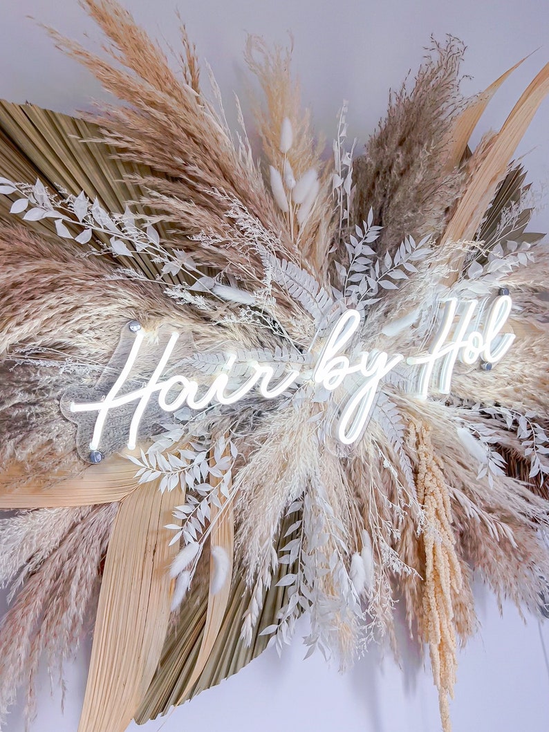 May include: A decorative wall hanging featuring dried pampas grass, palm leaves, and white floral accents. A neon sign that reads "Hair by Hol" is mounted in the center of the arrangement. The color palette includes beige, cream, and white.