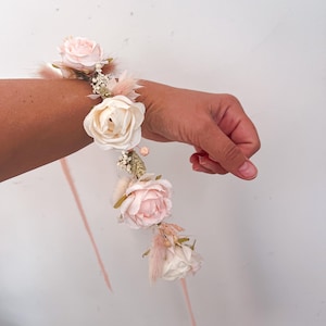 May include: A floral wrist corsage featuring light pink and cream-colored roses, accented with delicate feathers and small white flowers. The wristband is adorned with a matching ribbon, perfect for weddings or special events.