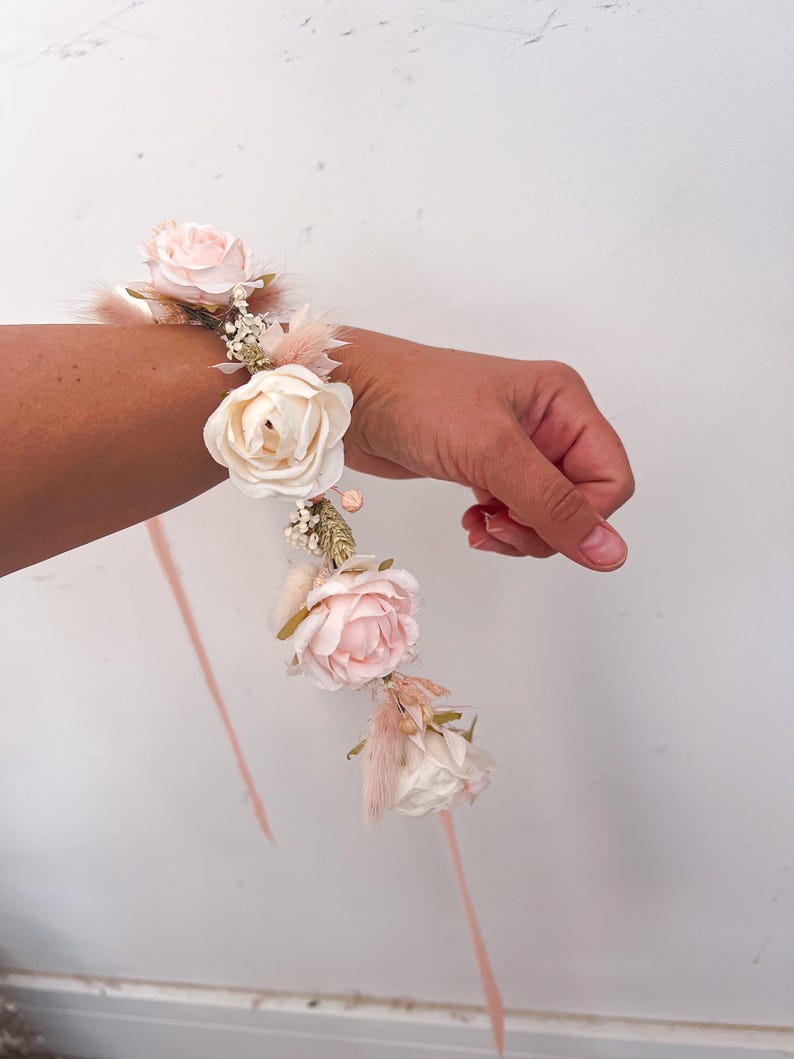 May include: A floral wrist corsage featuring light pink and cream-colored roses, accented with delicate feathers and small white flowers. The wristband is adorned with a matching ribbon, perfect for weddings or special events.