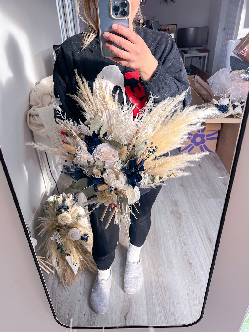 May include: A bouquet of dried flowers with white and cream coloured flowers, blue berries, and tan pampas grass.