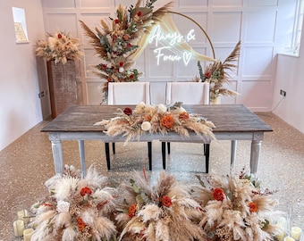 May include: Wedding reception decor featuring a wooden table with floral arrangements of pampas grass, roses, and greenery. A neon sign reads "Always & Forever." Additional floral arrangements and candles complete the scene.