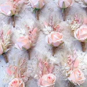 May include: A collection of blush pink rose boutonnieres with dried pampas grass and other delicate floral accents. Each boutonniere features a single rose, wrapped stems, and a pin for easy attachment. Perfect for weddings or special events.