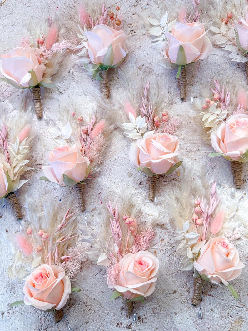 May include: A collection of blush pink rose boutonnieres with dried pampas grass and other delicate floral accents. Each boutonniere features a single rose, wrapped stems, and a pin for easy attachment. Perfect for weddings or special events.