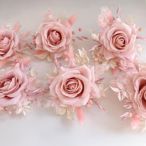 May include: A collection of blush pink roses with delicate, feathery accents and small, pearl-like beads. The roses are the focal point, surrounded by dried grasses and leaves in shades of pink and cream. The arrangement is set against a soft, neutral background.