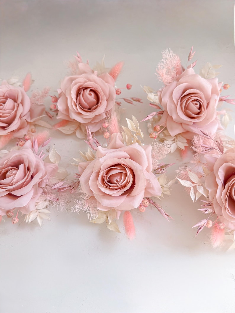 May include: A collection of blush pink roses with delicate, feathery accents and small, pearl-like beads. The roses are the focal point, surrounded by dried grasses and leaves in shades of pink and cream. The arrangement is set against a soft, neutral background.