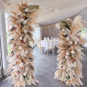May include: Two decorative floral arrangements featuring pampas grass, roses, and greenery. The arrangements are in beige, cream, and peach tones, suitable for a wedding or event. The background shows a dining area with tables and chairs.