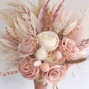 May include: A hand holding a bouquet of dried flowers. The bouquet features large, light pink roses, cream-colored roses, and various dried grasses and accents in shades of pink, cream, and beige. The arrangement is tightly packed and appears to be freshly made.