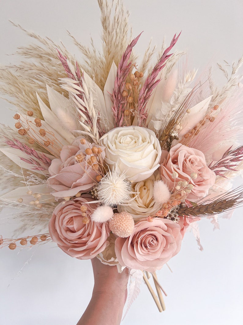 May include: A hand holding a bouquet of dried flowers. The bouquet features large, light pink roses, cream-colored roses, and various dried grasses and accents in shades of pink, cream, and beige. The arrangement is tightly packed and appears to be freshly made.
