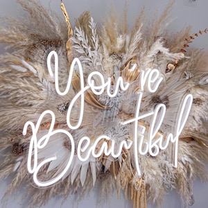 May include: White neon sign with the words "You're Beautiful" against a backdrop of dried pampas grass and other dried floral elements.