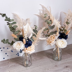 May include: Two small bouquets of dried flowers in clear glass vases. The bouquets feature cream and white roses, blue flowers, dried grasses, and pampas grass.