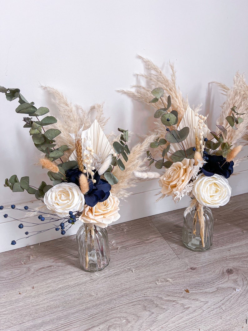 May include: Two small bouquets of dried flowers in clear glass vases. The bouquets feature cream and white roses, blue flowers, dried grasses, and pampas grass.
