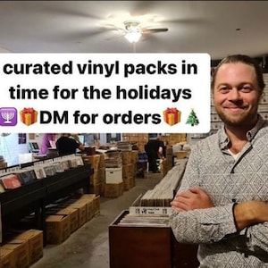 May include: A man standing in a record store with shelves full of vinyl records. The text on the sign reads "curated vinyl packs in time for the holidays DM for orders".