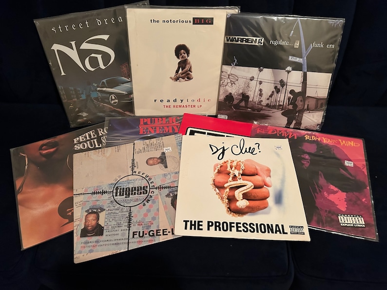 May include: A collection of nine vinyl records, including albums by Nas, The Notorious B.I.G., Warren G, Public Enemy, Fu-Gee-La, DJ Clue?, and Redman. The records are all in protective plastic sleeves and are arranged in a stack on a dark blue surface.