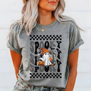 May include: Gray t-shirt with a black and white checkered pattern at the top and bottom. The shirt features a cartoon ghost wearing a beanie hat and holding a pumpkin and a cup of coffee. The text "Spooky" is repeated in a retro font.