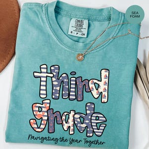 May include: Seafoam green t-shirt with the words "Third Grade" in a nautical-themed design. The shirt features anchors, sailboats, and fish patterns. The text "Navigating the Year Together" is printed below the main text. A gold necklace is draped over the shirt.