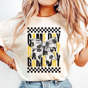 May include: A white t-shirt with a black and yellow checkered pattern and the text "GAME DAY" repeated three times. A black and white bow with a soccer ball pattern is in the center of the design.