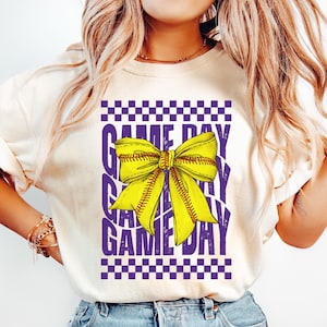 May include: A white t-shirt with a purple and yellow graphic design. The graphic features a yellow bow with a baseball stitch pattern and the text "GAME DAY" repeated in a purple, distressed font.