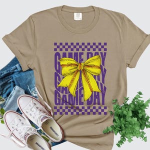 May include: Khaki t-shirt with a purple and yellow graphic that says "Game Day" with a yellow bow in the center.
