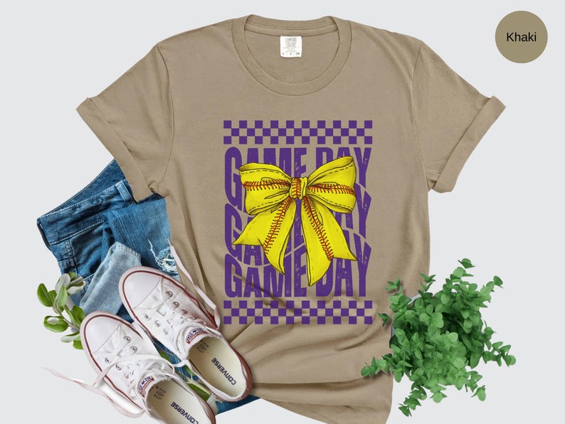 May include: Khaki t-shirt with a purple and yellow graphic that says "Game Day" with a yellow bow in the center.
