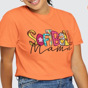K&ouml;nnte beinhalten: Ein leuchtend oranges T-Shirt mit den Worten "Softball Mama" in bunten, gemusterten Buchstaben. Die Buchstaben sind mit Sternen, Herzen und N&auml;hten verziert.
