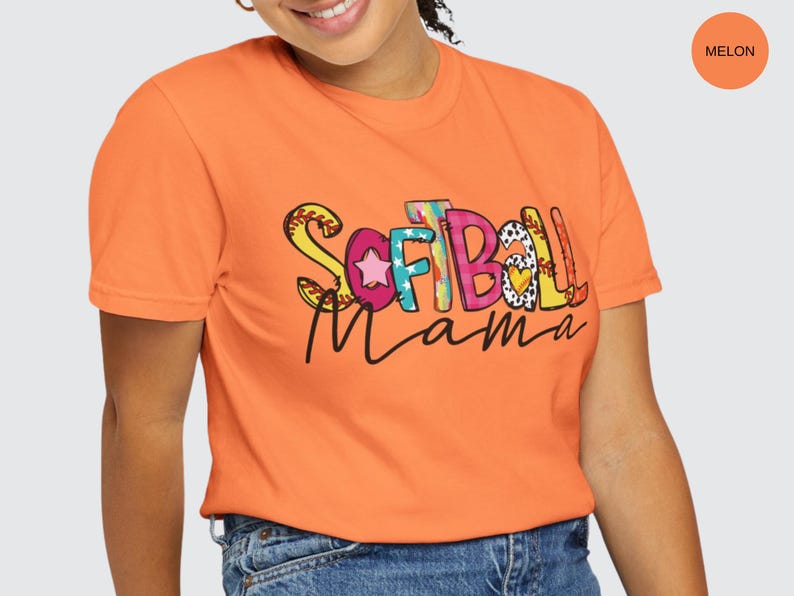 K&ouml;nnte beinhalten: Ein leuchtend oranges T-Shirt mit den Worten "Softball Mama" in bunten, gemusterten Buchstaben. Die Buchstaben sind mit Sternen, Herzen und N&auml;hten verziert.