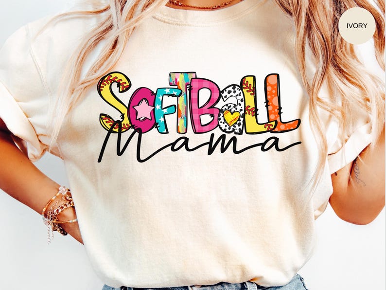 K&ouml;nnte beinhalten: Ein wei&szlig;es T-Shirt mit dem Schriftzug "Softball Mama" in bunten, verspielten Buchstaben. Die Buchstaben sind mit Baseballn&auml;hten, Sternen und Herzen verziert.