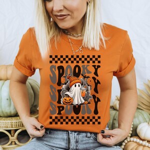 May include: A burnt orange t-shirt with a graphic of a ghost holding a pumpkin and a drink. The text "Spooky" is repeated in a checkered pattern on the shirt.