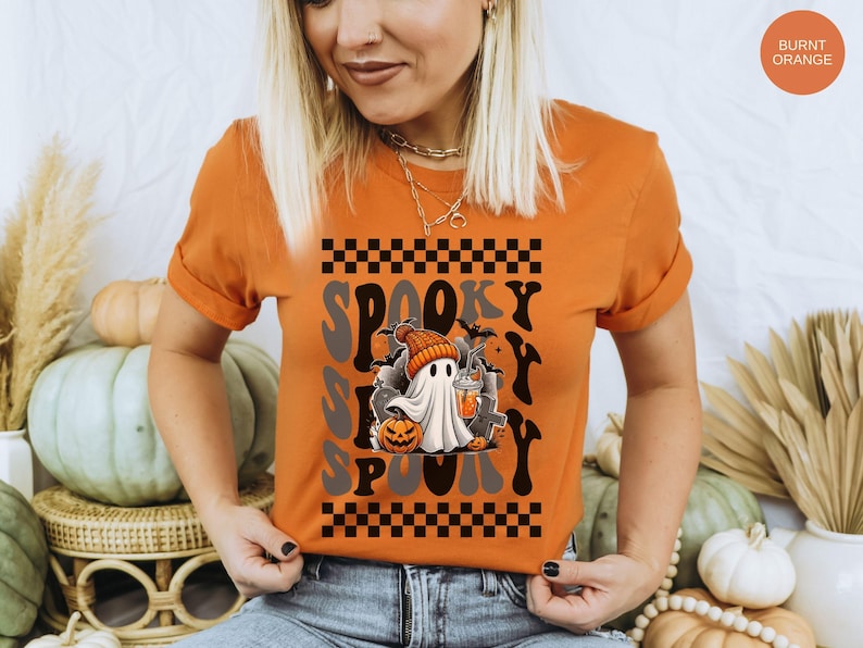 May include: A burnt orange t-shirt with a graphic of a ghost holding a pumpkin and a drink. The text "Spooky" is repeated in a checkered pattern on the shirt.