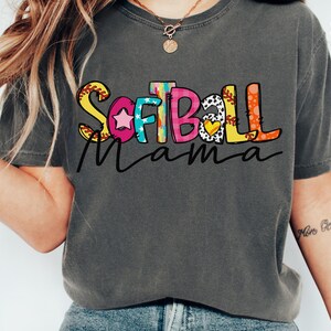 K&ouml;nnte beinhalten: Ein graues T-Shirt mit dem Text "Softball Mama" in bunten Buchstaben. Die Buchstaben sind mit einem Baseball, Sternen und anderen Designs verziert.