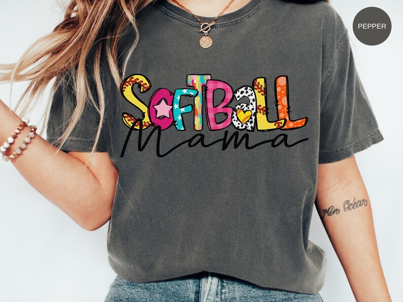 K&ouml;nnte beinhalten: Ein graues T-Shirt mit dem Text "Softball Mama" in bunten Buchstaben. Die Buchstaben sind mit einem Baseball, Sternen und anderen Designs verziert.