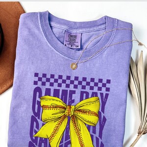 May include: Purple t-shirt with a yellow and white softball bow and the text "Game Day" in purple. The shirt is a comfortable, relaxed fit.