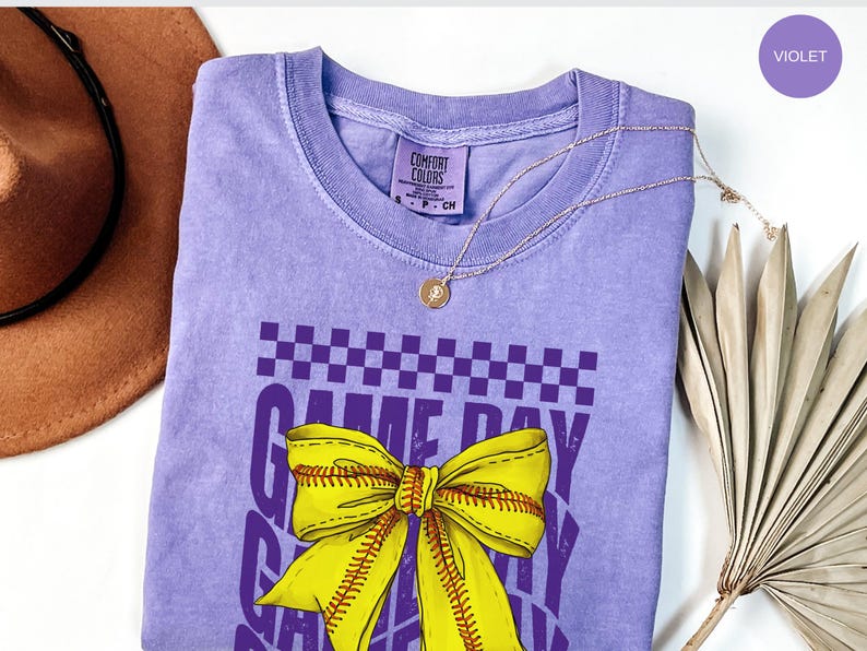 May include: Purple t-shirt with a yellow and white softball bow and the text "Game Day" in purple. The shirt is a comfortable, relaxed fit.