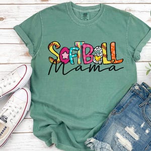 K&ouml;nnte beinhalten: Ein hellgr&uuml;nes T-Shirt mit dem Text "Softball Mama" in bunten Buchstaben mit einer Softball-Grafik. Das Shirt hat einen Used-Look und ist aus einem weichen, bequemen Material gefertigt.