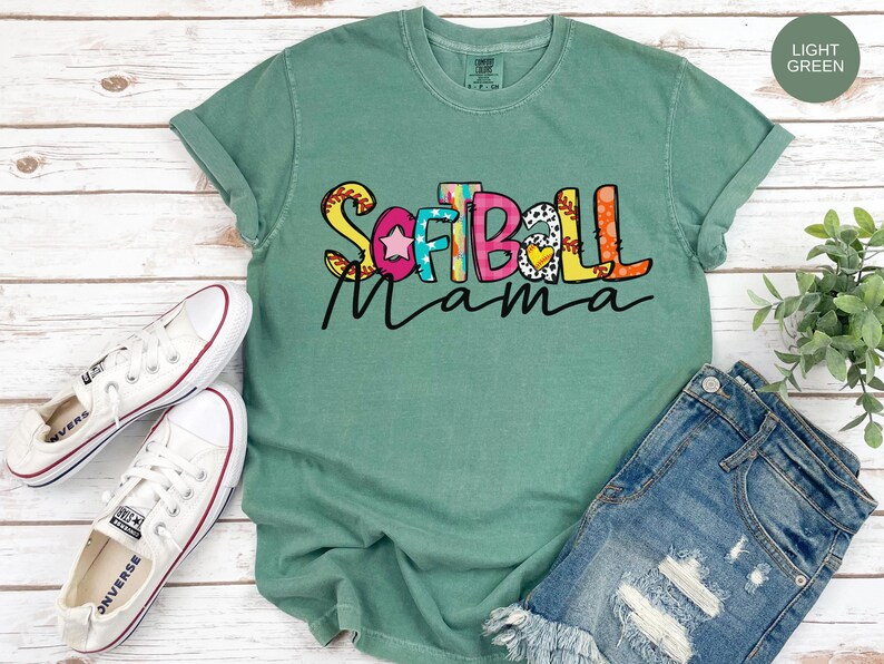 K&ouml;nnte beinhalten: Ein hellgr&uuml;nes T-Shirt mit dem Text "Softball Mama" in bunten Buchstaben mit einer Softball-Grafik. Das Shirt hat einen Used-Look und ist aus einem weichen, bequemen Material gefertigt.